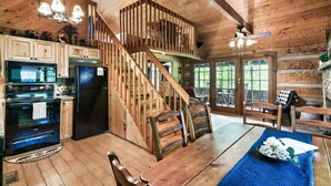 L’intérieur d’un chalet rustique comprend un escalier en bois, un coin repas avec une table et des chaises, une cuisine équipée d’appareils modernes et une grande fenêtre offrant une vue sur l’extérieur.