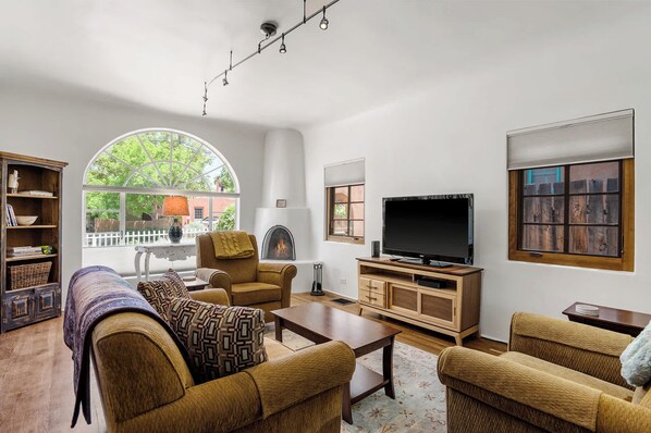 A living room with a fireplace, a television, and a large window overlooking a white fence and greenery. - Right on the Plaza next to O'keeffe Museum (Santa Fe)