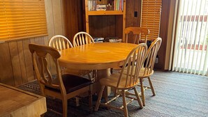 Een houten eettafel met zes stoelen, een boekenkast vol boeken en decoratieve objecten, en een wandklok.