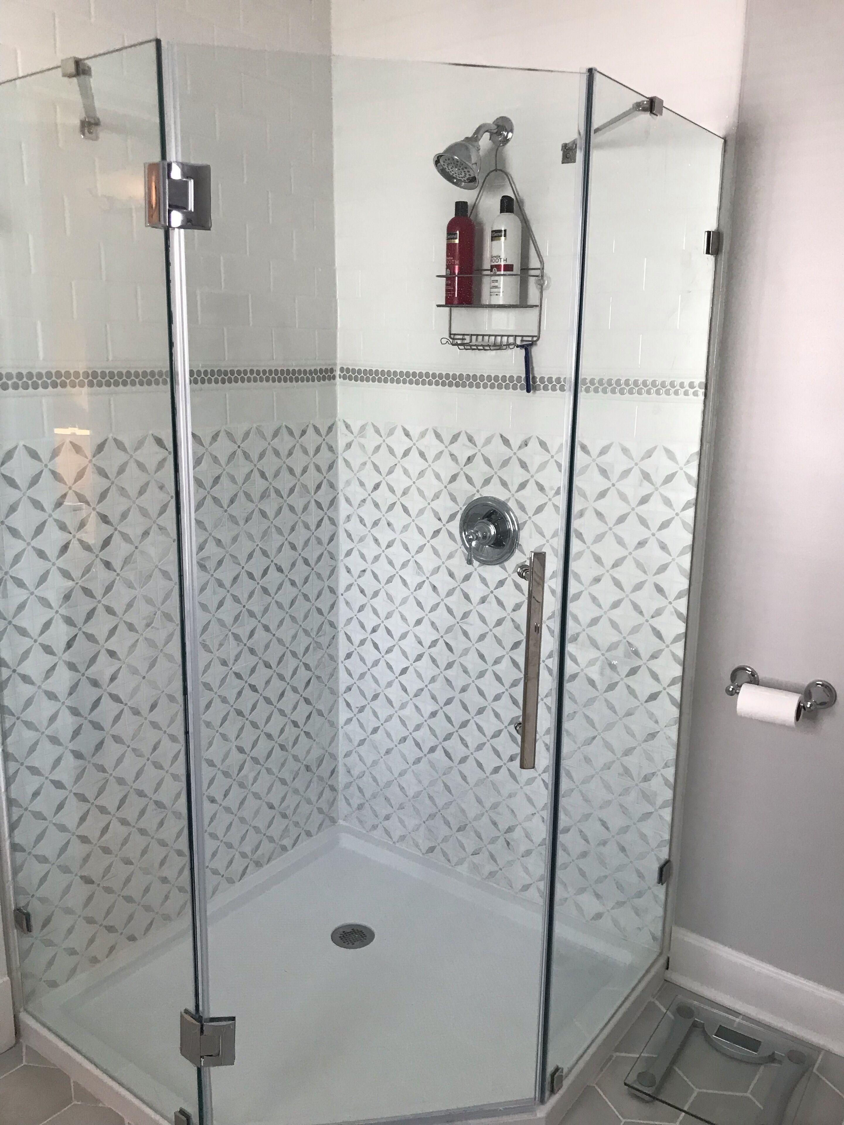 Eine gläserne Dusche mit geometrischem Fliesenspiegel an den Wänden und einem Metall-Duschkopf.