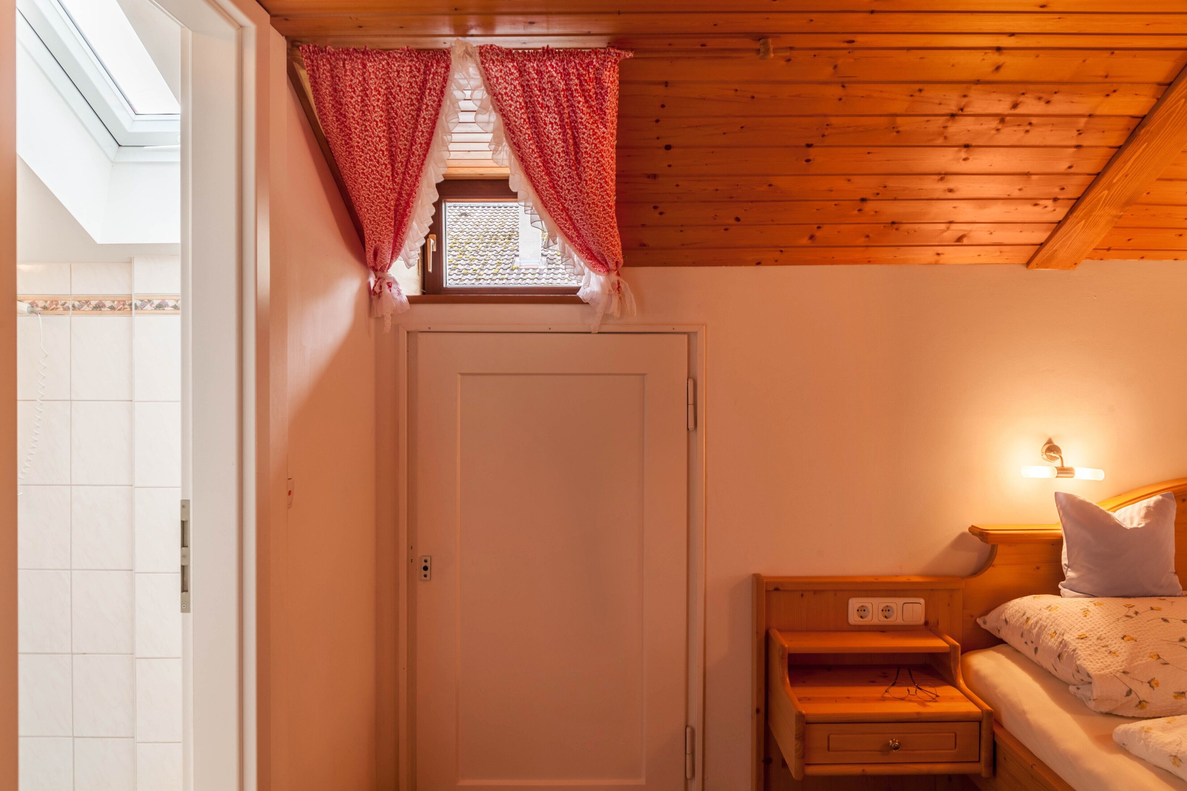 Ein Zimmer mit Holzboden, einer weißen Tür, einem kleinen Fenster mit roten Vorhängen, einem Nachttisch mit Lampe und einem Bett mit Kissen.