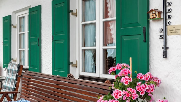 Ein weißes Gebäude mit grünen Fensterläden, einer Holzbank und rosa Blumen im Vordergrund.