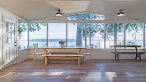 Amplia habitación con un gran ventanal con vistas a un cuerpo de agua, un banco de madera y una mesa con sillas.