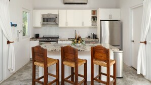 Een keuken met witte kastjes, een roestvrijstalen koelkast, een magnetron en een spoelbak. Er staan drie houten barkrukken met gevlochten zittingen.