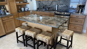 Une cuisine dotée d’un plan de travail en granit, de meubles en bois, d’une cuisinière en acier inoxydable et de quatre tabourets en osier.
