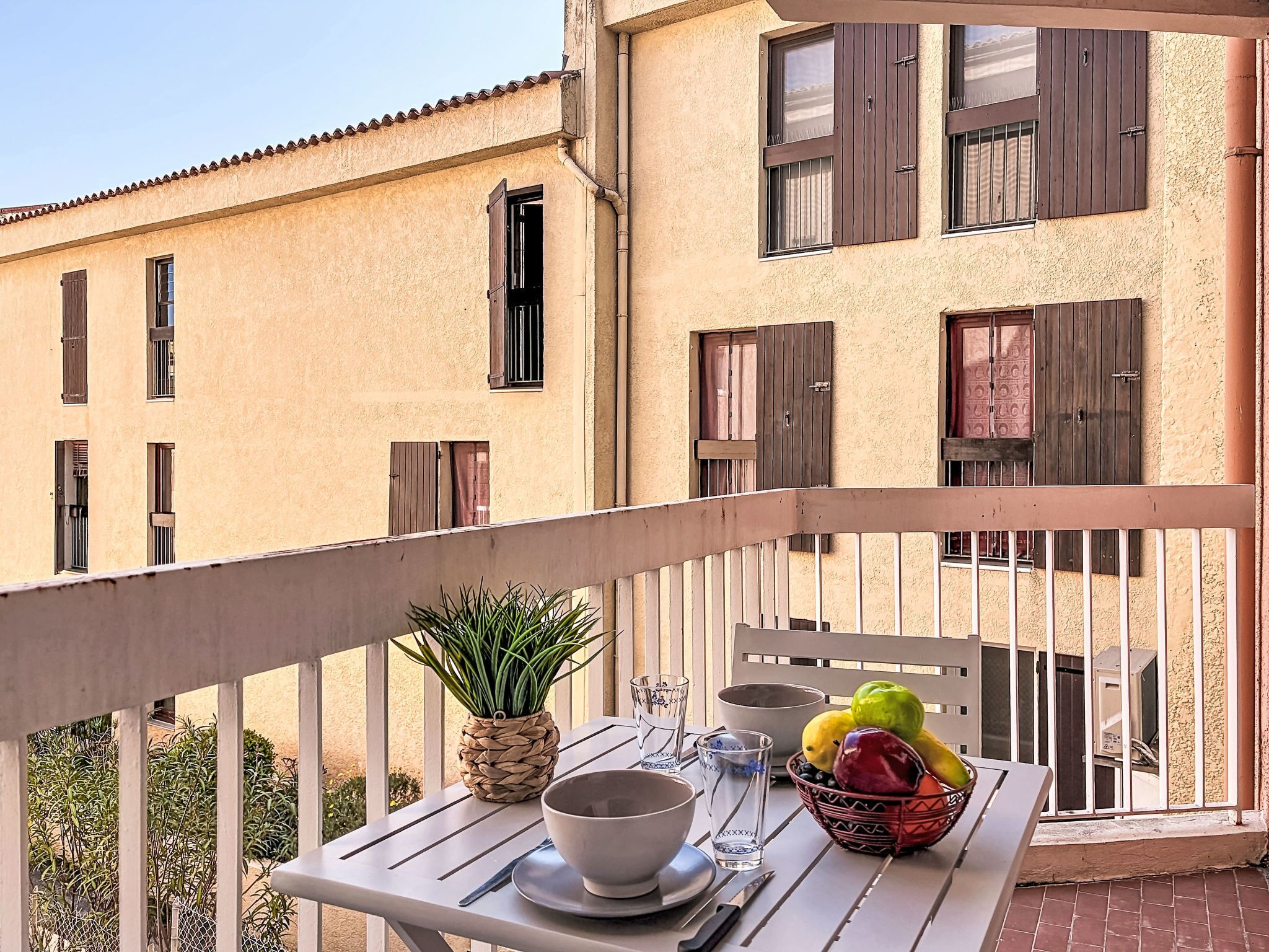 Un balcon avec une table dressée pour le dîner, une plante en pot et un saladier rempli de fruits.
