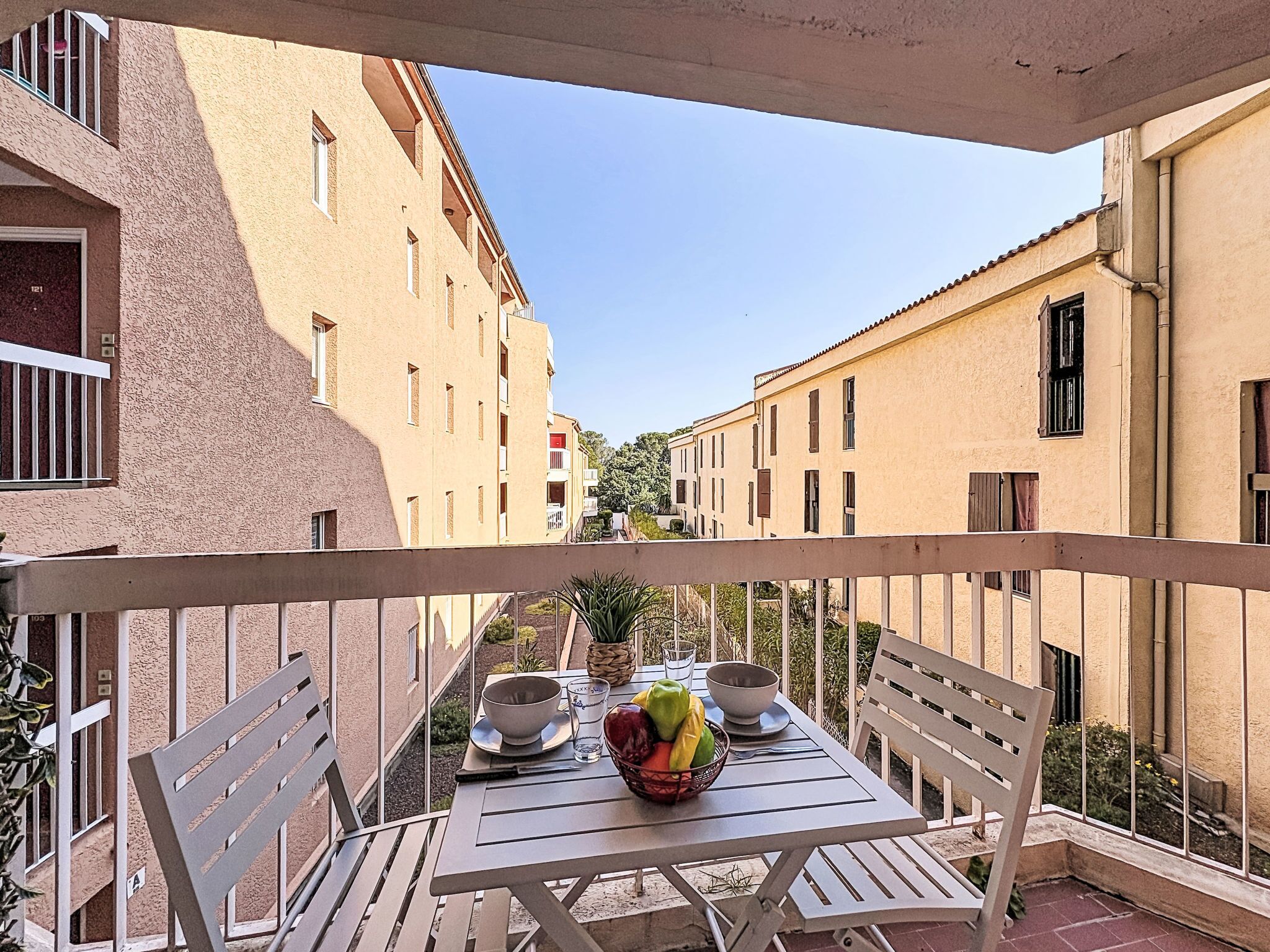 A balcony with a table set for two, a bowl of fruit, and a view of adjacent buildings.