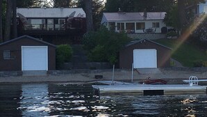 Une propriété située au bord de l’eau, comprenant deux bâtiments de type garage, un quai avec un bateau et, en arrière-plan, une rangée d’arbres imposants.
