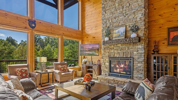 A cozy living room with a stone fireplace, leather sofas, and a large window overlooking a forest. - Secluded 4BRMountain Retreat:Stunning Views, Pool Table, Arcade, HotTub, FirePit (Sevierville)
