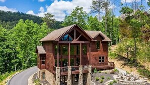 A large wooden cabin with a balcony, surrounded by a stone foundation and a landscaped garden with a fire pit. - Secluded 4BRMountain Retreat:Stunning Views, Pool Table, Arcade, HotTub, FirePit (Sevierville)