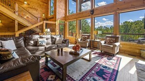 A spacious living area with a large wooden staircase, leather sofas, a coffee table, and large windows offering a view of the outdoors. - Secluded 4BRMountain Retreat:Stunning Views, Pool Table, Arcade, HotTub, FirePit (Sevierville)