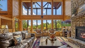 A spacious living room with a leather sofa, armchairs, a coffee table, and a stone fireplace. Large windows offer a view of the outdoors. - Secluded 4BRMountain Retreat:Stunning Views, Pool Table, Arcade, HotTub, FirePit (Sevierville)