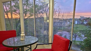 Une pièce avec une table en verre et deux chaises rouges, de grandes fenêtres donnant sur un parc et des immeubles.