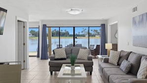 A spacious living room with a large window overlooking a body of water, a sofa, a coffee table, and a lamp. - Lakeside Sunset Retreat | Ground Floor | 3 En Suites | Optional Saddlebrook Resort Amenities (Wesley Chapel)