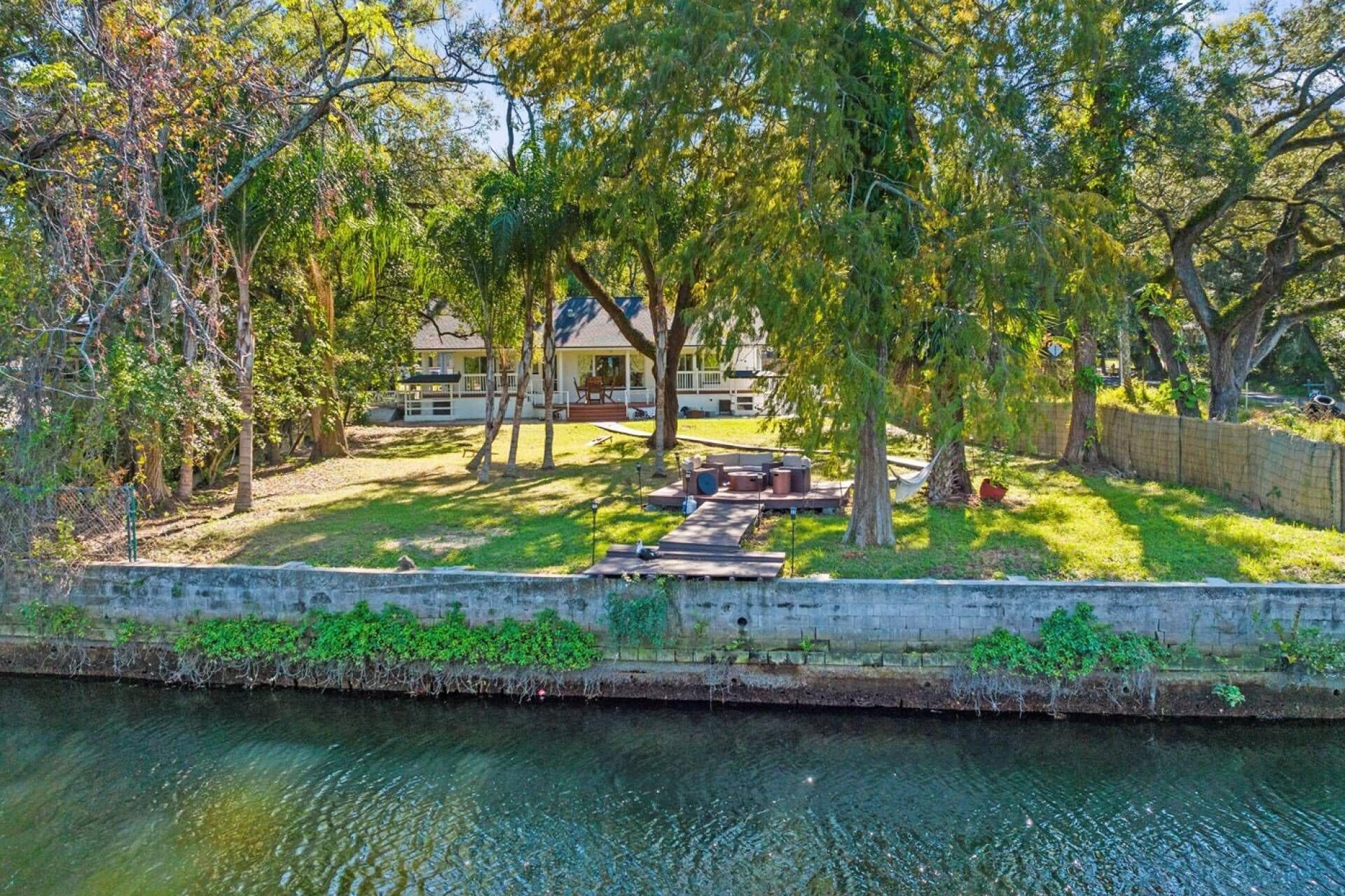 A house with a deck and outdoor furniture, surrounded by trees and a body of water.