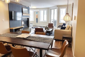 Un salon moderne avec une cheminée, un coin repas avec une longue table et des chaises, et un espace détente avec un canapé et des fauteuils.