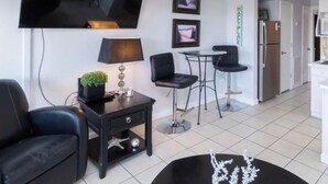 A modern living room with a flat-screen TV, a small table with a lamp, two black chairs, and a decorative plant. - Beach. View. Suite. 1 br. 1 ba. Sleeps 5 (Hilton Head Island)