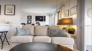 A modern living room with a beige sectional sofa, a black coffee table, and a TV mounted on the wall. There are framed pictures on the wall and a lamp on a side table. - Beach. View. Suite. 1 br. 1 ba. Sleeps 5 (Hilton Head Island)