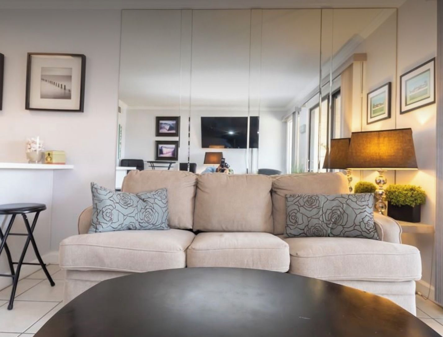 Ein modernes Wohnzimmer mit einem beigefarbenen Ecksofa, einem schwarzen Couchtisch und einem an der Wand montierten Fernseher. An den Wänden befinden sich Bilder in Rahmen und auf einem Beistelltisch steht eine Lampe.