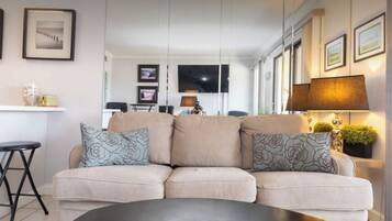 Ein modernes Wohnzimmer mit einem beigefarbenen Ecksofa, einem schwarzen Couchtisch und einem an der Wand montierten Fernseher. An den Wänden befinden sich Bilder in Rahmen und auf einem Beistelltisch steht eine Lampe.