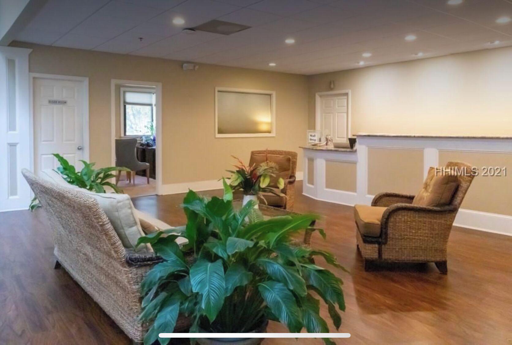 A spacious living room with a large sofa, two armchairs, and several potted plants. There are two doors, one labeled "Nurse Room" and another with a sign that reads "Mail Room." The room has a high ceiling with recessed lighting and a fireplace.