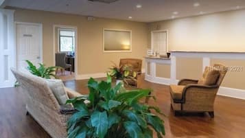 A spacious living room with a large sofa, two armchairs, and several potted plants. There are two doors, one labeled "Nurse Room" and another with a sign that reads "Mail Room." The room has a high ceiling with recessed lighting and a fireplace.