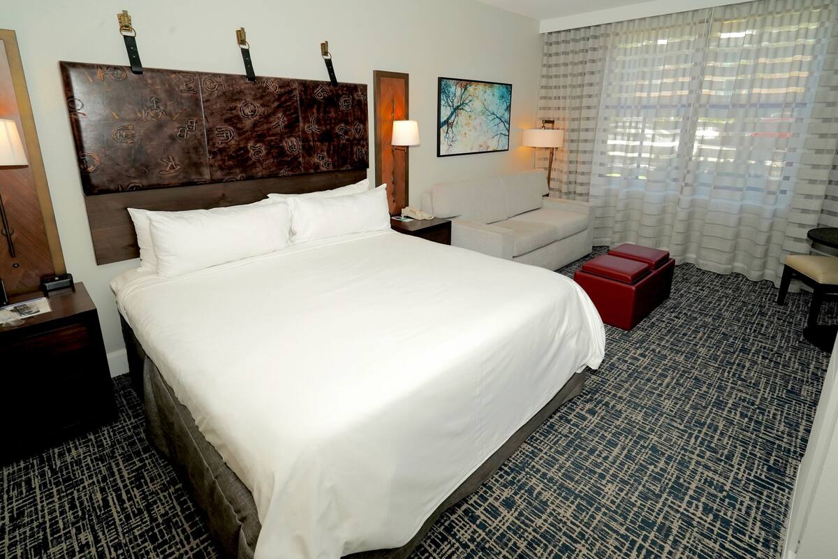 A hotel room with a large bed, a sofa, a red ottoman, a desk, and a headboard with a unique design.