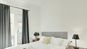 Ein ordentlich bezogenes Bett mit weißen Leinen, ein Nachttisch mit Lampe und Blick auf einen Balkon durch ein Fenster mit grauen Vorhängen.