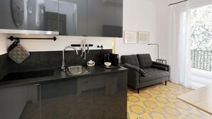 Et moderne køkken med en sort bordplade, stålvask og komfur i sort. Der er en grå sofa og et lille bord i det tilstødende opholdsområde.