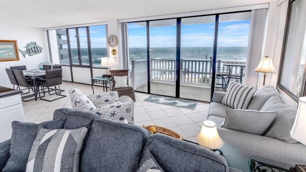 A spacious living area with a large balcony overlooking the ocean, featuring a dining table, seating area, and a view of the sea.