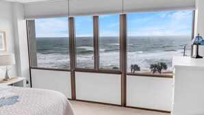 A bedroom with a large window overlooking the ocean, a bed, a nightstand, and a lamp.