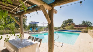 An outdoor pool area with a table and chairs under a wooden pergola, surrounded by greenery and a clear sky. - Charming villa with shared pool (Issac)