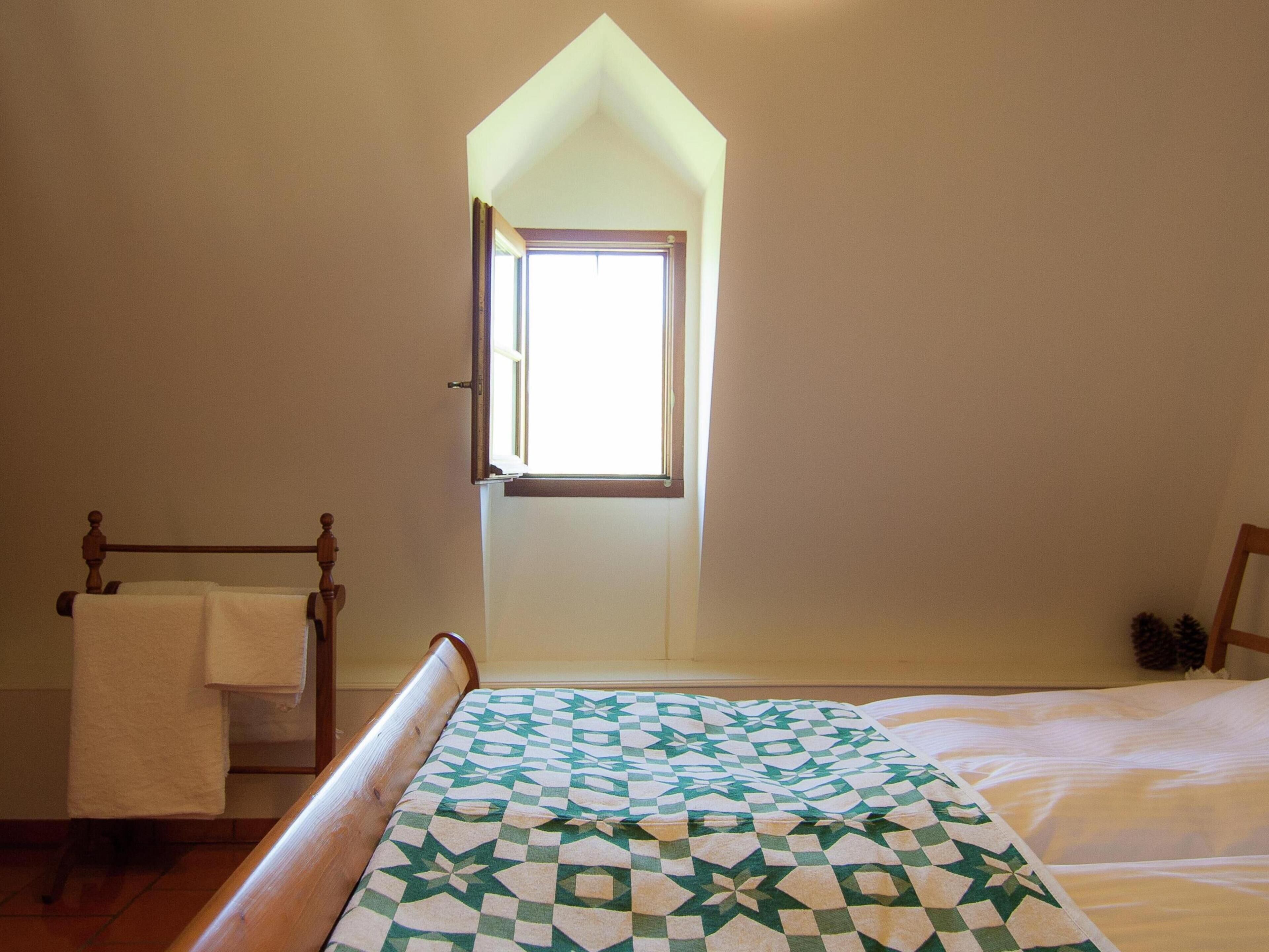 Ein Schlafzimmer mit schrägem Dach, einem Fenster, einem Bett mit gemustertem Überwurf, einem Holz-Kopfschild und einem Holzstuhl.