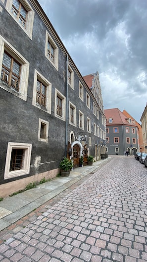 Una calle empedrada flanqueada por edificios históricos, incluyendo un teatro y un edificio con un letrero que dice 'Wirtshaus'.