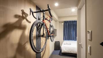 Quarto de hotel com cama, bicicleta presa na parede, mala e um alto-falante.