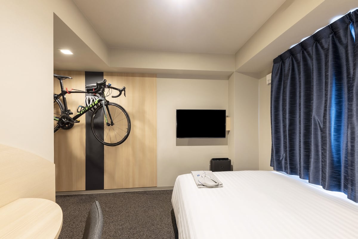 A hotel room with a bed, a TV mounted on the wall, a bicycle hanging on the wall, and a window with curtains.