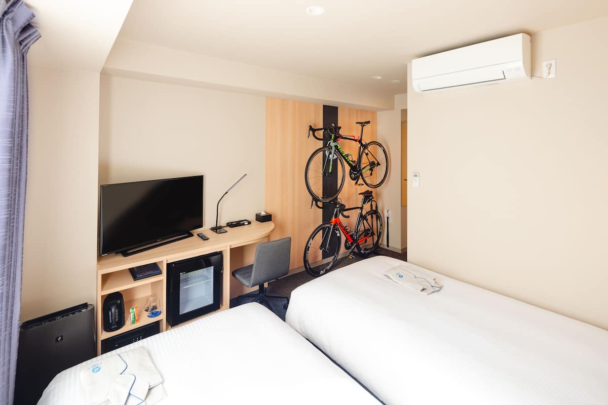 A hotel room with two beds, a TV, a desk with a computer, a chair, and a bicycle mounted on the wall.