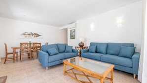 Una sala de estar con dos sofás azules, una mesa de centro de vidrio, una mesa de comedor de madera con sillas, y un cuadro en la pared.