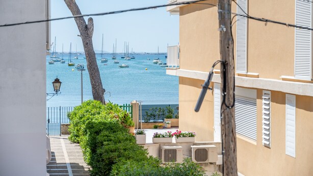 Vista costera con barcos, un balcón con plantas en macetas y un edificio con contraventanas.