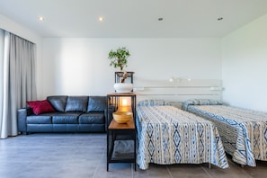 A modern hotel room with a sofa, a bedside table with a lamp, a patterned bed, and a potted plant.