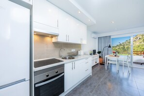 A modern kitchen with white cabinets, a stainless steel sink, and an oven. There is a dining area with a white table and chairs, and a view of a green landscape through large windows. - Comfortable Studio with Pool, Near Alcudia Beach (Alcudia)