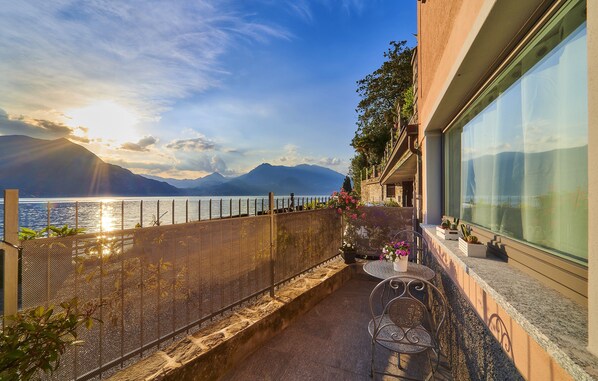 A balcony with a view of a lake and mountains, a table with chairs, and potted plants.
