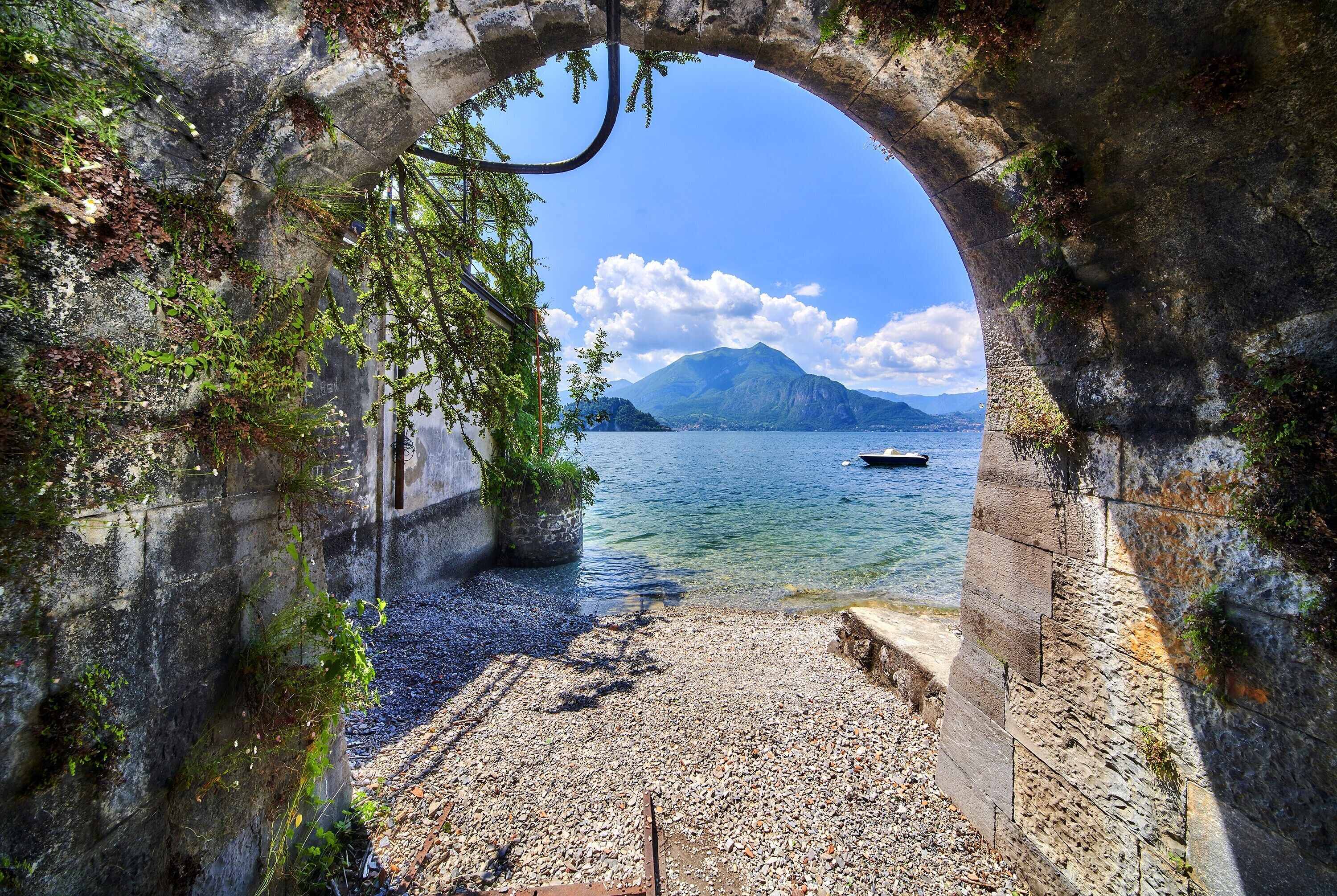 Eine Bogenkonstruktion aus Stein, die zu einem Kiesstrand führt, mit freiem Blick auf einen Berg und ein Gewässer.