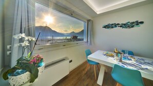 Une pièce comprenant une table à manger, des chaises, une vue sur la montagne et la mer, un élément décoratif mural et une fenêtre avec des rideaux.