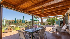A covered patio with a wooden ceiling, outdoor furniture, and a view of a garden.