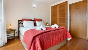 Un lit bien fait, recouvert d’une couverture rouge, une lampe de chevet et une armoire en bois.