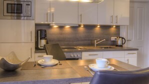 A modern kitchen with a built-in microwave, a coffee maker, and a kettle on the countertop. There are two white cups on a table with a placemat. - Villa Namenlos - Holiday apartment 5 (Börgerende-Rethwisch)