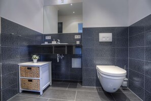 A modern bathroom with dark blue tiles, a white sink, a toilet, and wicker baskets on a cabinet. - Villa Namenlos - Vacation Apartment 5 (Börgerende-Rethwisch)