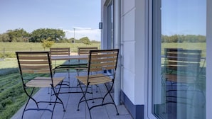 A patio with four wooden chairs and a table, a glass door, and a view of a grassy field and trees. - Villa Zeitlos - Holiday apartment 1 (Börgerende-Rethwisch)
