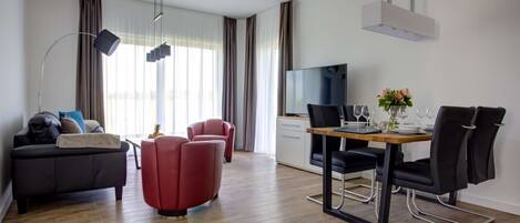 A modern living room with a sofa, red chairs, a dining table with chairs, a TV, and a wooden floor.